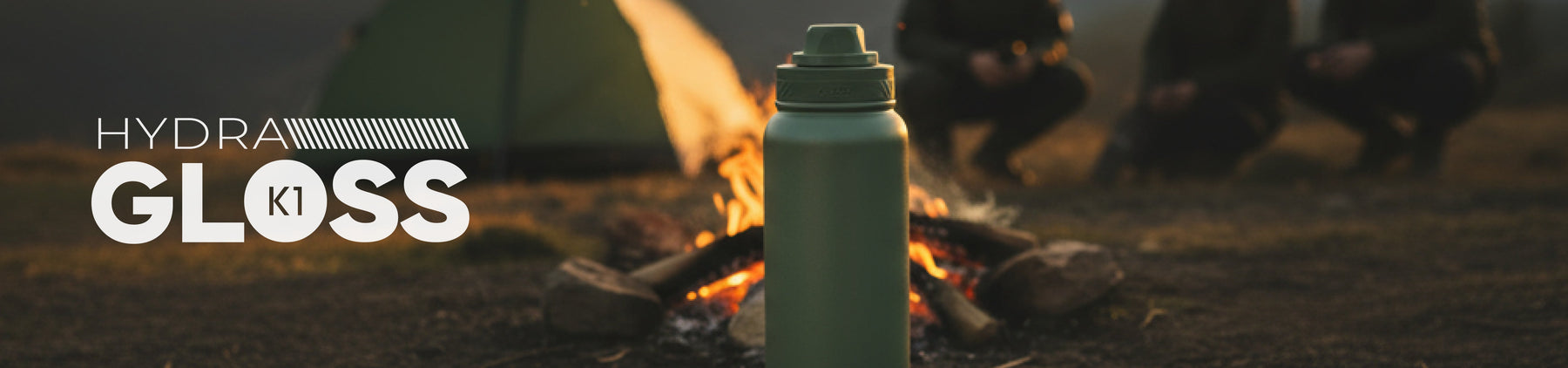 Garrafa térmica verde colocada sobre o chão de terra, em frente a uma fogueira acesa. Duas pessoas ao fundo, com clima de acampamento ao entardecer. Texto: Hydra Gloss K1.